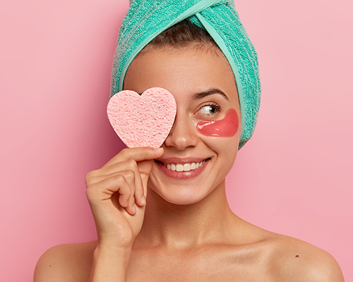 Mujer con una toalla verde en la cabeza y antiojeras se tapa un ojo con una esponja en forma de coraz&oacute;n mientras sonr&iacute;e